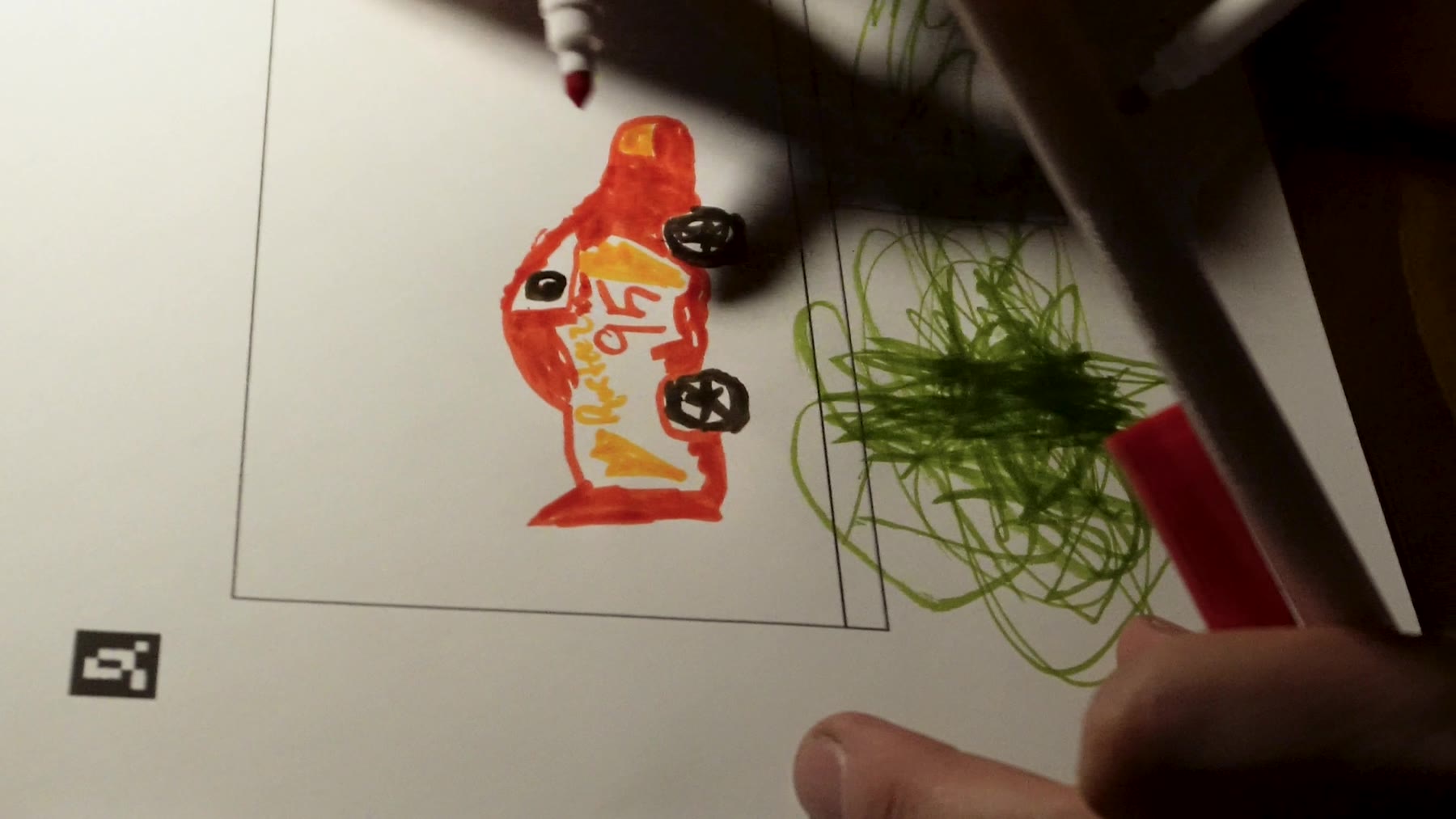 Macro shot of hands coloring a car-shaped billboard page in orange and black marker, with a small square fiducial marker printed in the corner of the page