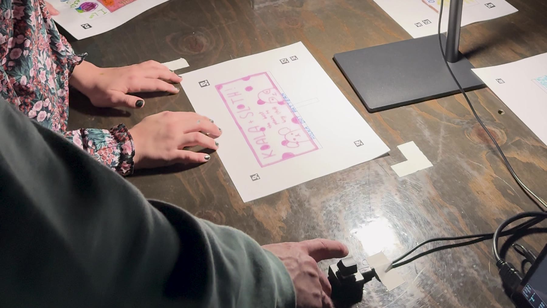 Two people at a wooden table: one holds a finished pink 'Koala + Sloth' drawing flat under a small overhead light/camera rig while a hand presses a small black button device
