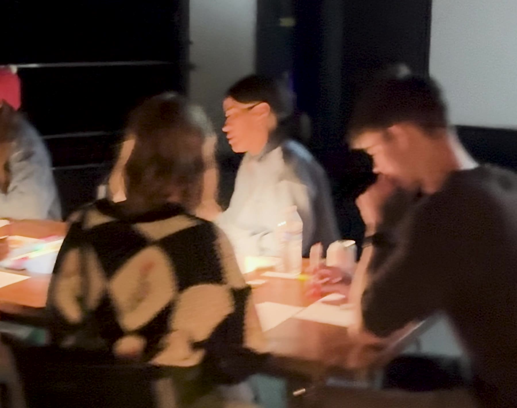 Adults seated around a table strewn with colored drawings and water bottles, mid-conversation, lit by small warm table lamps in a dark room