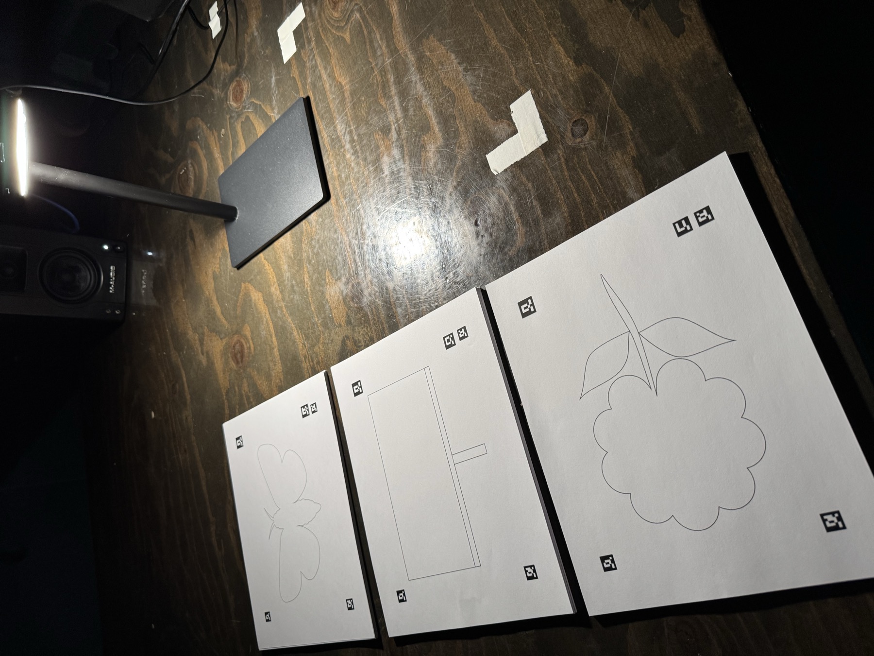 Three blank coloring pages laid out on the wooden table under a lamp: a butterfly, a billboard and a flower, each with four small printed fiducial markers near its corners