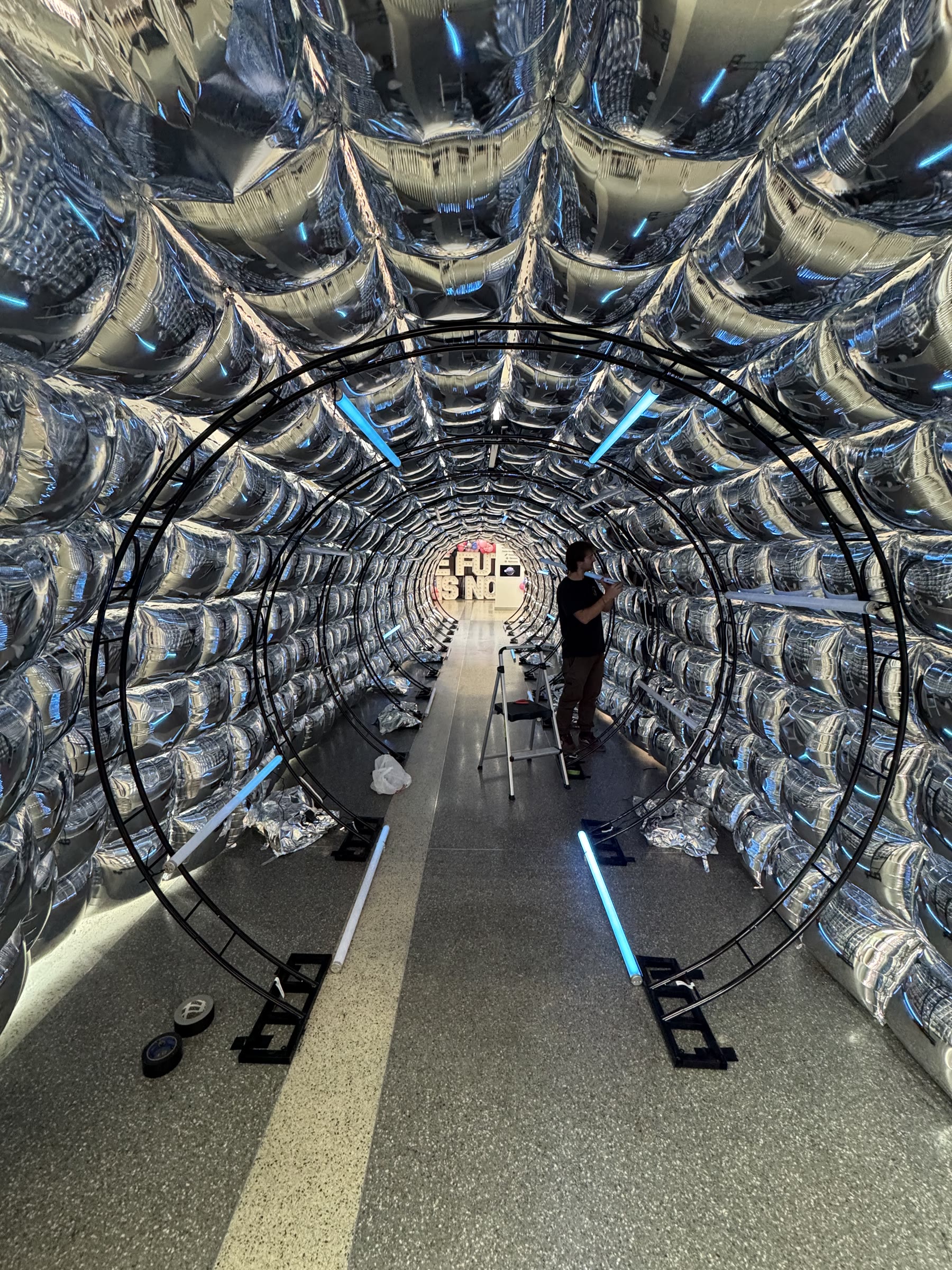 Looking down the length of the mylar balloon tunnel during setup, the black truss hoops and a few Astera tubes lit blue, a crew member on a ladder near the far end