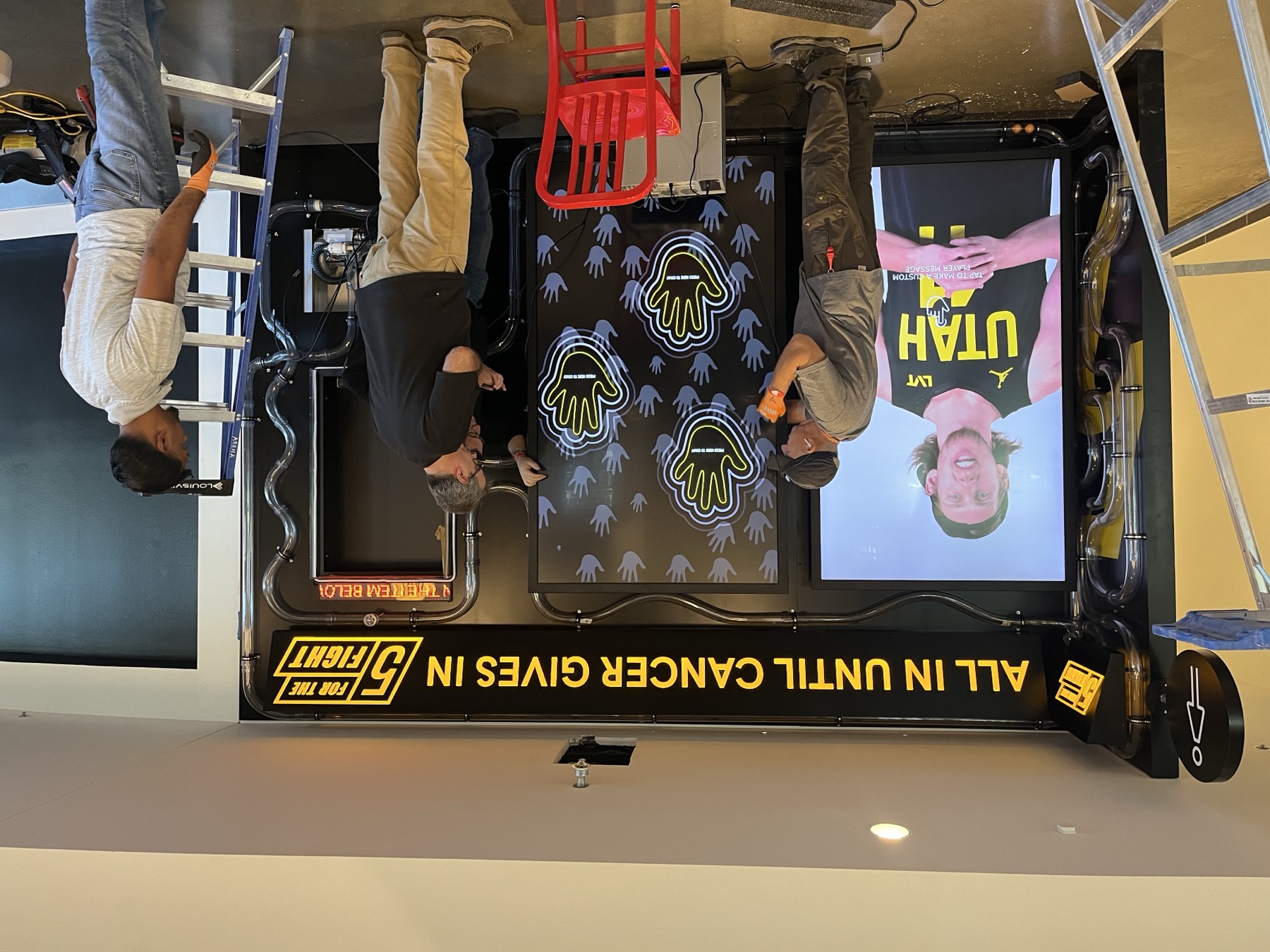 Install day at the Delta Center: crew on ladders fitting the two portrait screens into the black wall, the player video already playing on the left screen and the handprint prompts on the right