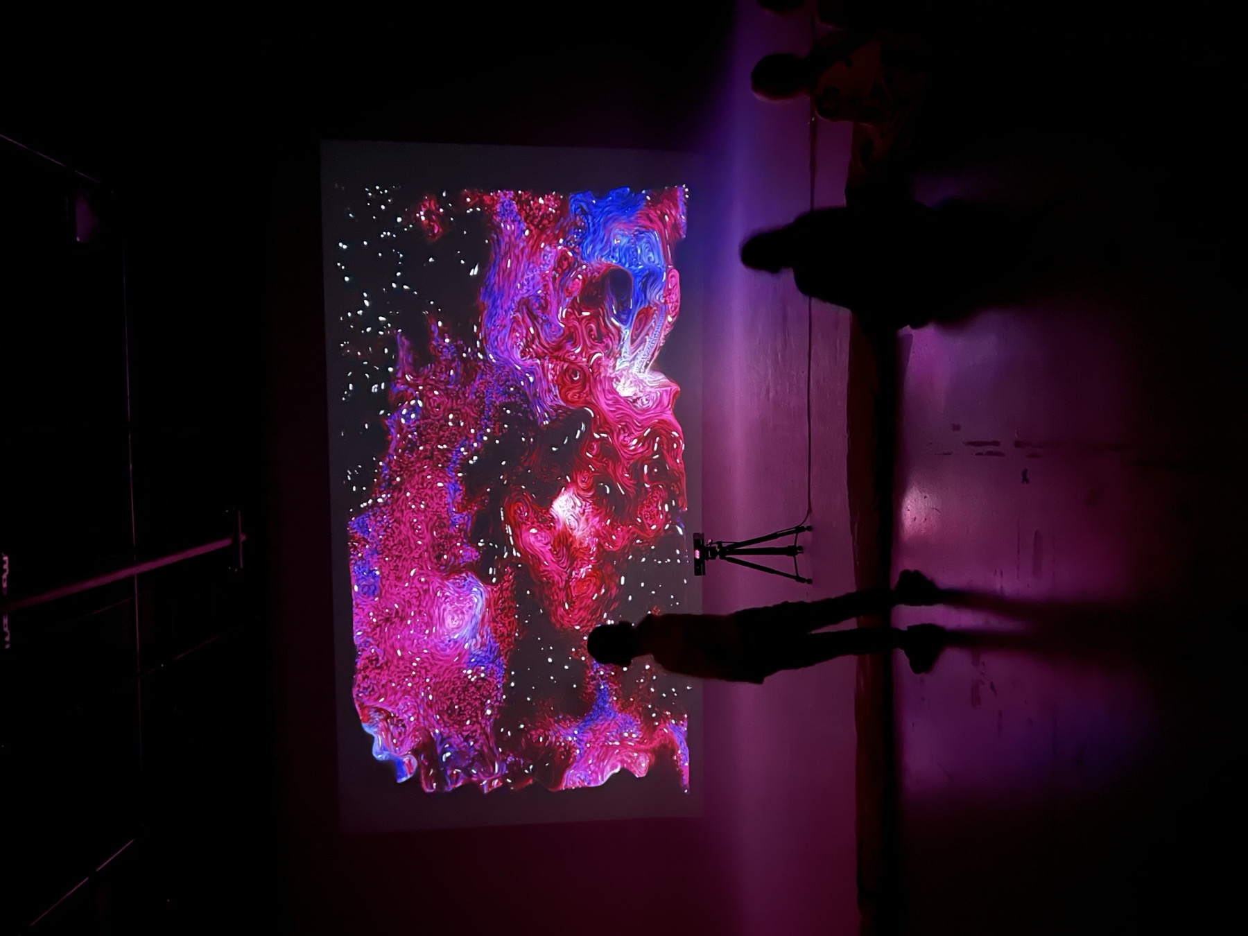 Wide shot of the full projection wall showing a red, magenta and blue swirling particle field, one silhouetted figure standing in front near a tripod sensor and two more seated at the base of the wall