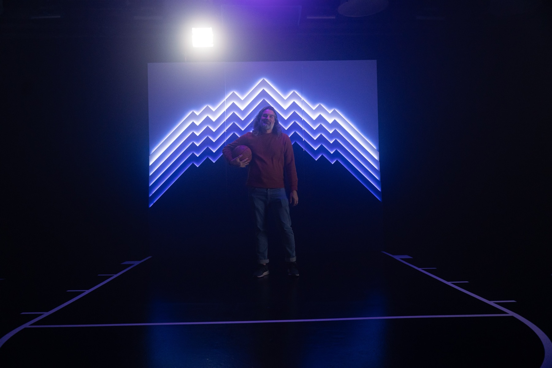 A man holding a basketball poses in front of the illuminated mountain sculpture on a marked court in a darkened studio