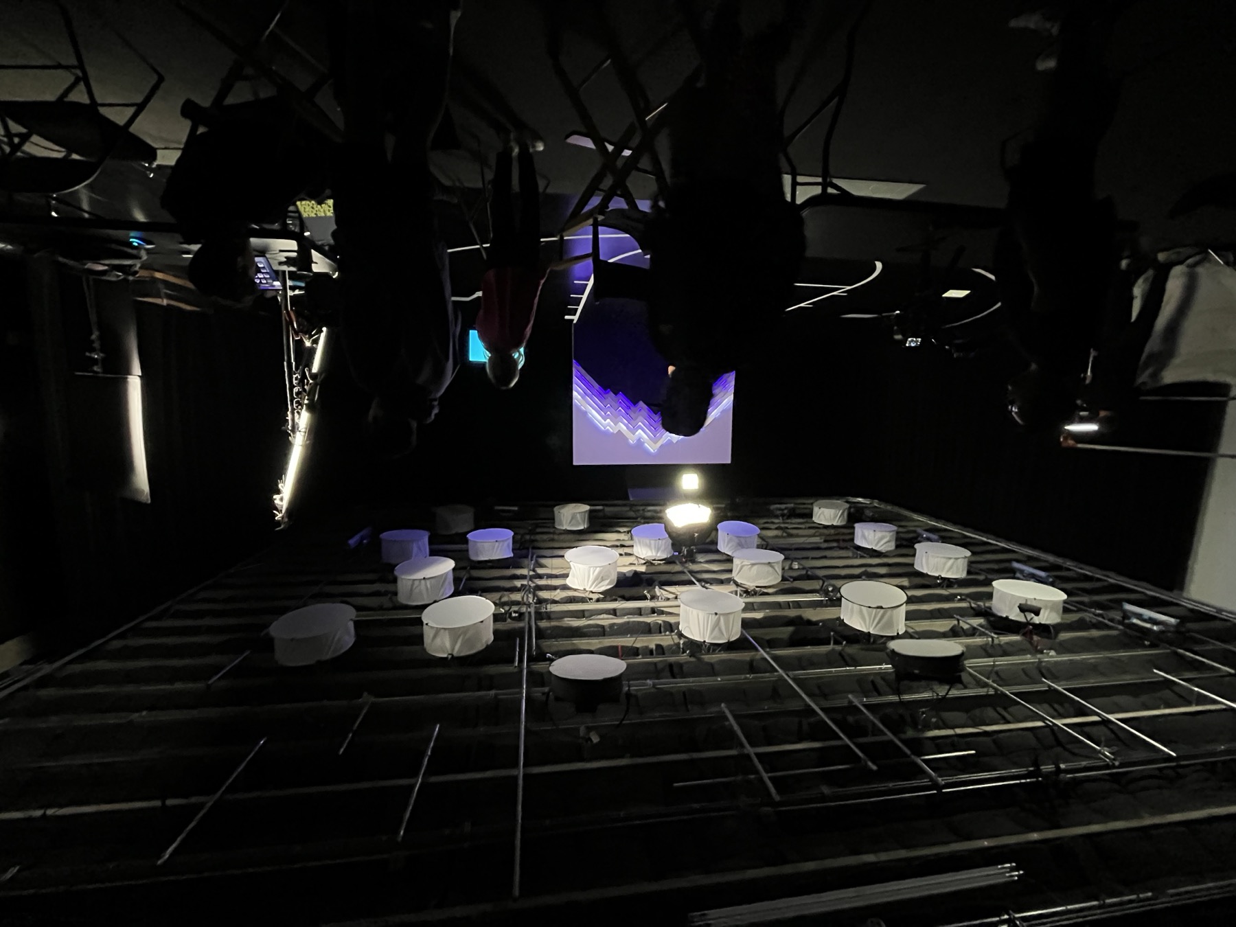 The video shoot in a dark studio: the mountain wall glowing white and violet at center under a grid of softbox lights, crew silhouetted in the foreground and a court line taped on the floor