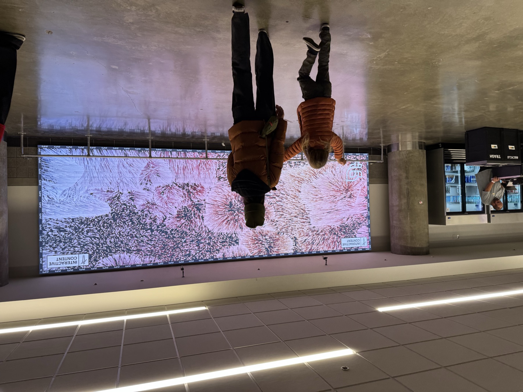 An adult and a small child, seen from behind, walking toward the lit wall in the daytime concourse
