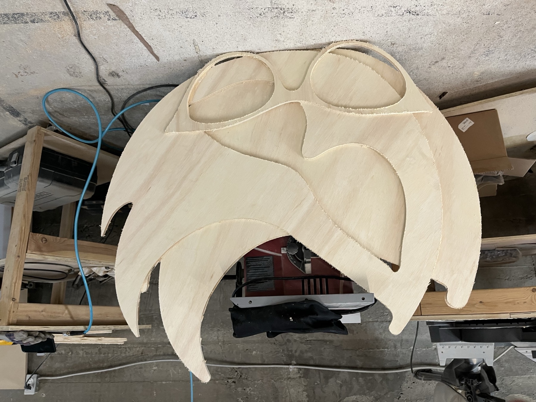 Stack of CNC-cut plywood mask shapes in the shop, the cat-eye glasses outline visible in each one