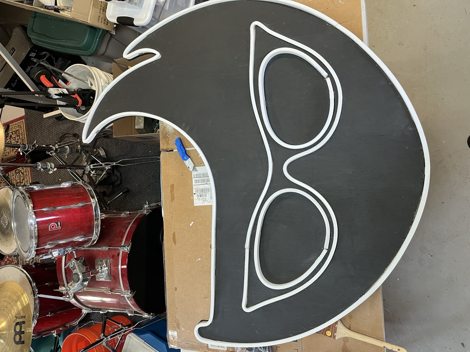 The mask sign painted black with the white outline and glasses drawn on, leaning against a drum kit