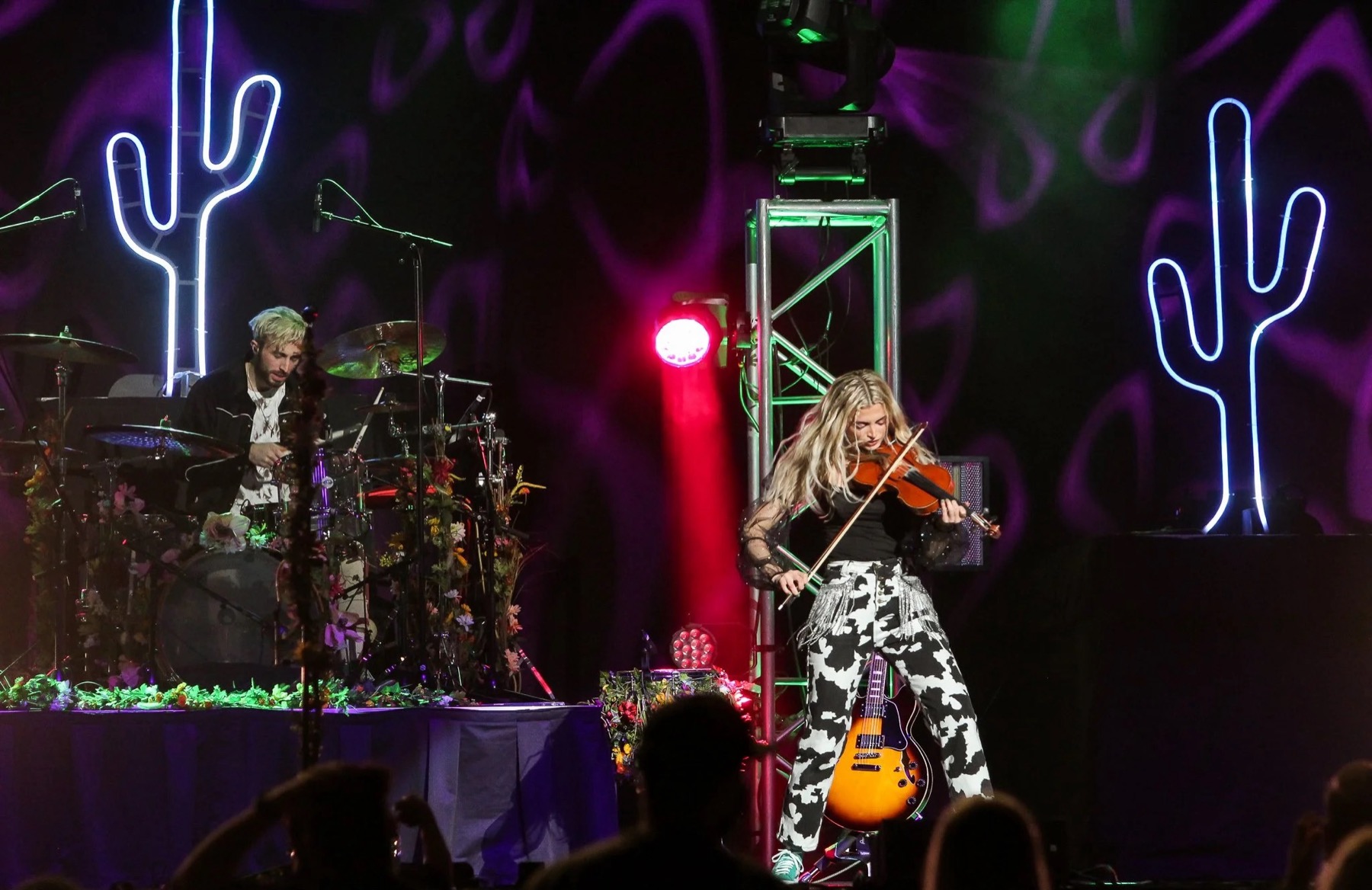 Two neon cactus fixtures glowing blue-white on either side of the stage, a drummer at left and a violinist at center under purple and green lighting