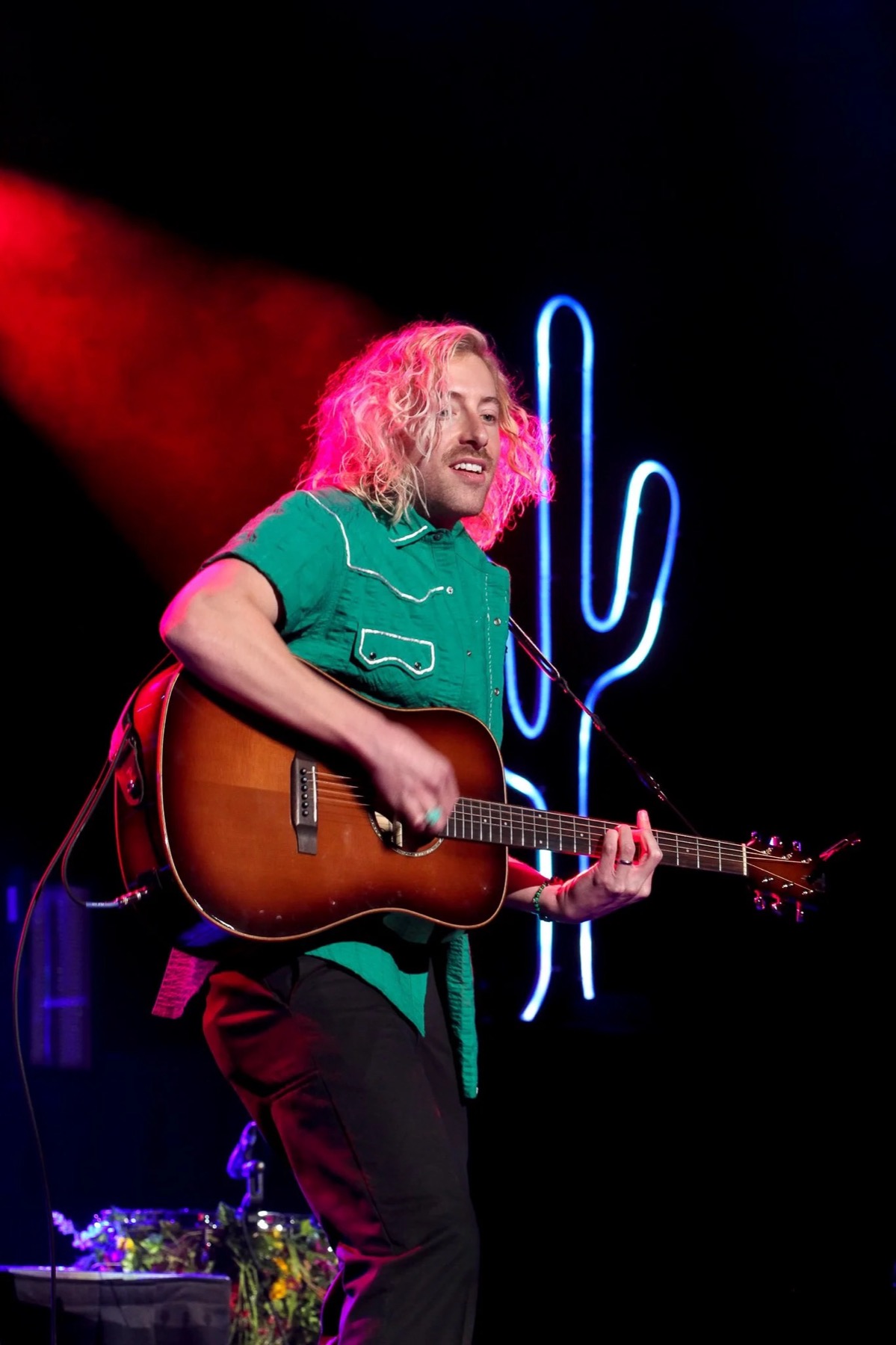Guitarist singing under a red spotlight with a neon cactus glowing blue behind him