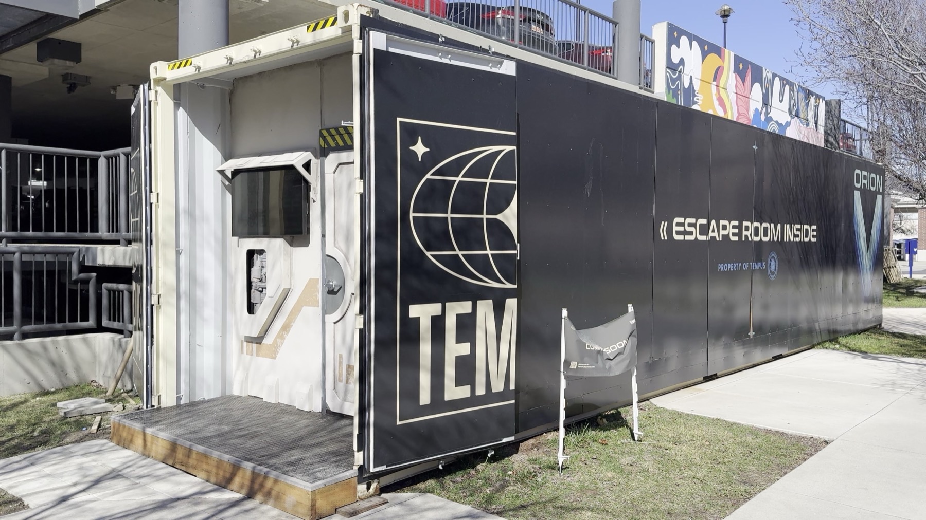 Exterior of a black shipping container with gold TEM globe logo and white text reading ESCAPE ROOM INSIDE, parked outside a building with a colorful mural above