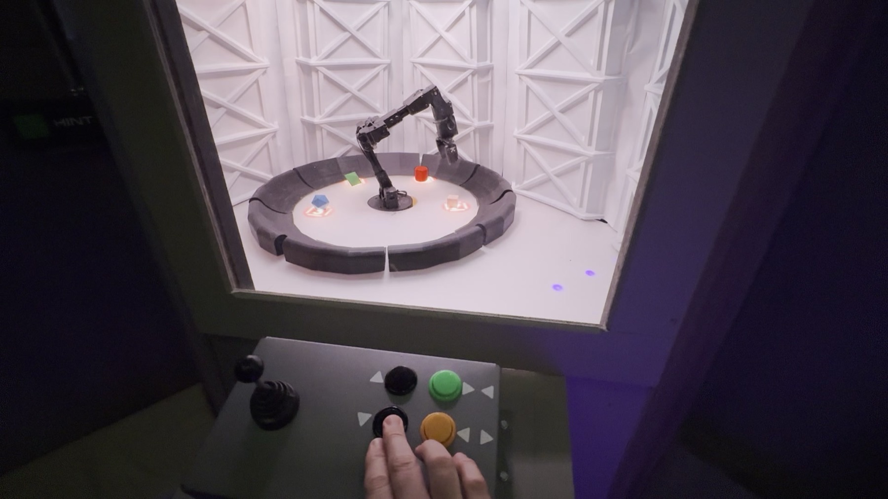 A small robotic arm inside a lit white chamber with colored geometric objects on a circular platform, controlled by a joystick and colored arcade buttons in the foreground with a player's finger on one button
