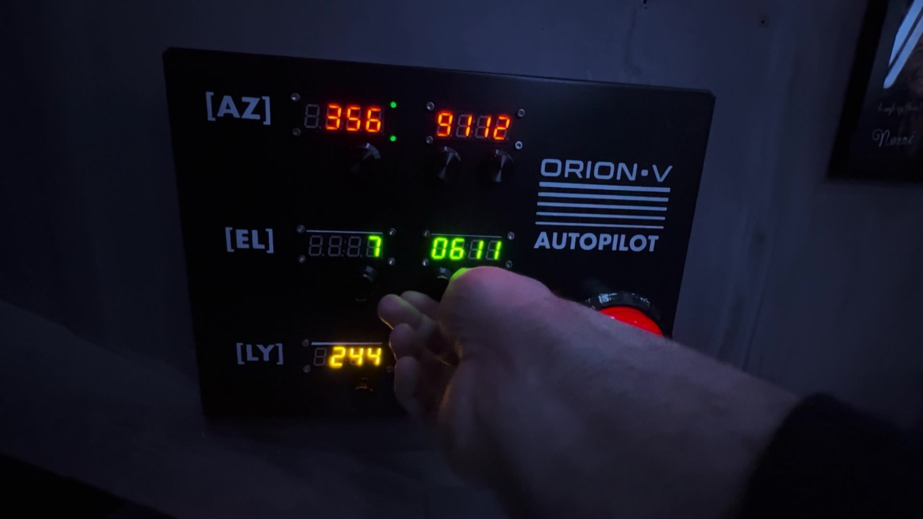 Close-up of a black control panel labeled ORION-V AUTOPILOT with lit AZ/EL/LY digital readouts in red, green and yellow, three rotary dials, a red button, and a player's hand adjusting a dial