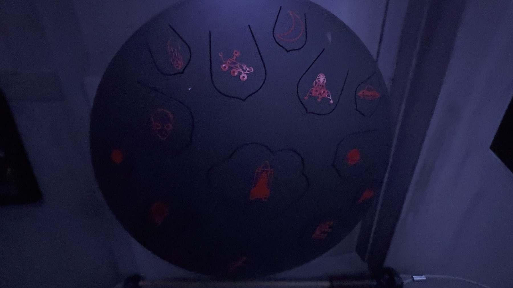 Close-up of a tongue drum (steel handpan-style percussion instrument) with hand-painted glyphs in each note slot: a skull, a rover, a crescent moon, a rocket, a UFO, and others, lit in dim blue light