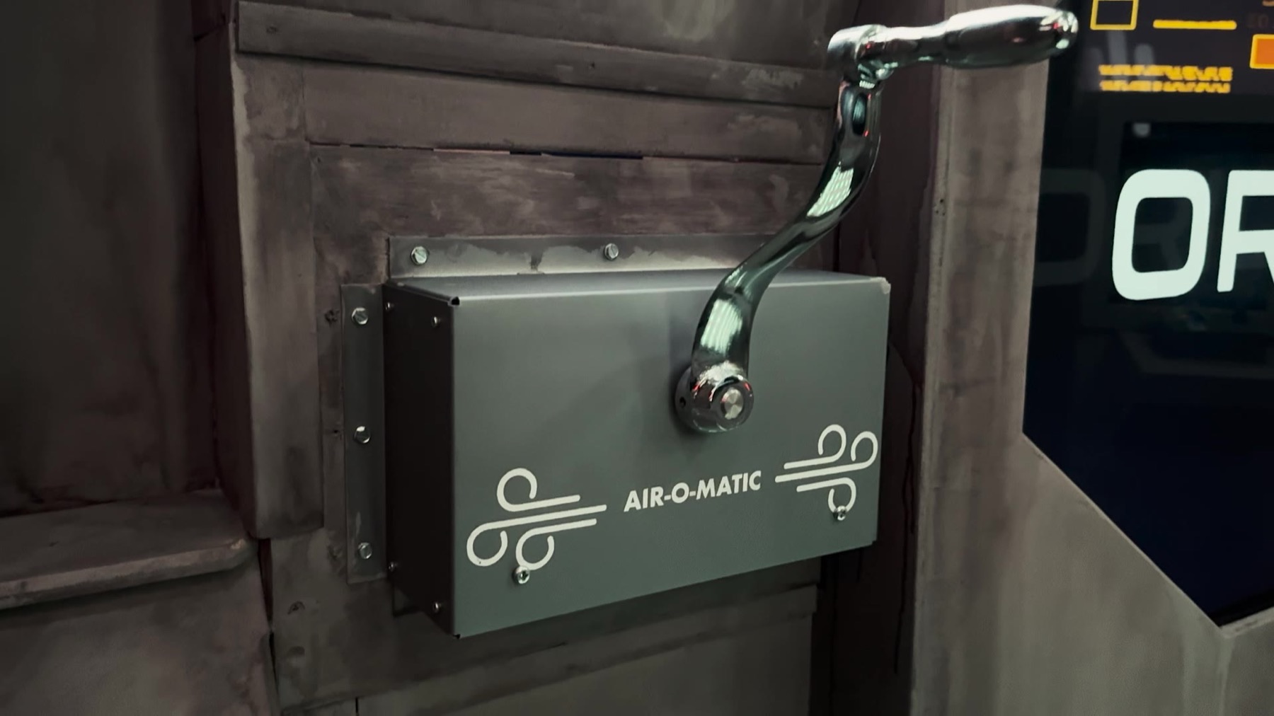 A gray metal box labeled AIR-O-MATIC with a wind/vent icon and a chrome hand-crank lever mounted on a distressed wood-paneled wall