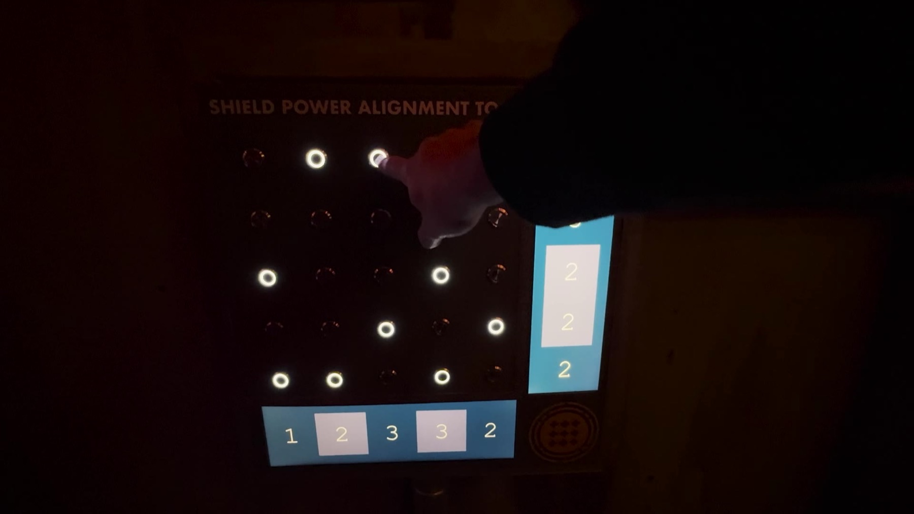 A dark panel reading SHIELD POWER ALIGNMENT TOOL with a 5x5 grid of lit and unlit ring buttons, a blue readout column showing repeating numbers, and a player's hand pressing one of the lit buttons
