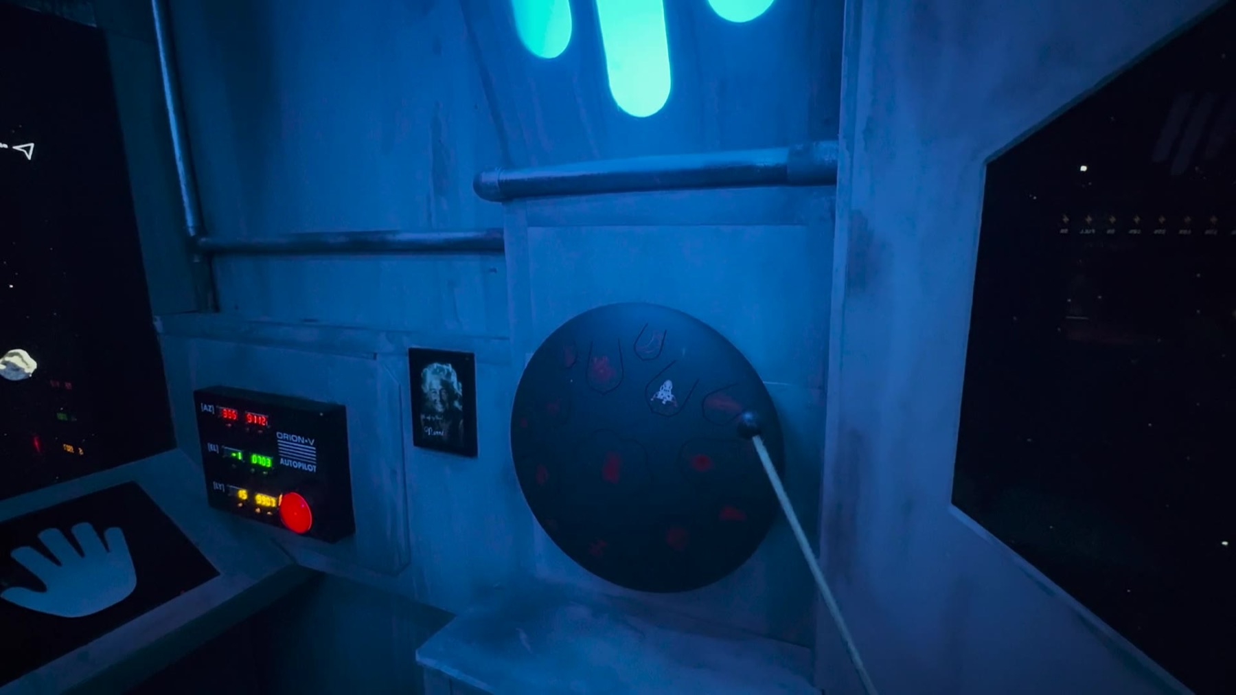 Atmospheric interior view in blue lighting showing the ORION-V AUTOPILOT panel, a framed photo, the tongue-drum instrument with its mallet, and a round porthole window with a starfield graphic