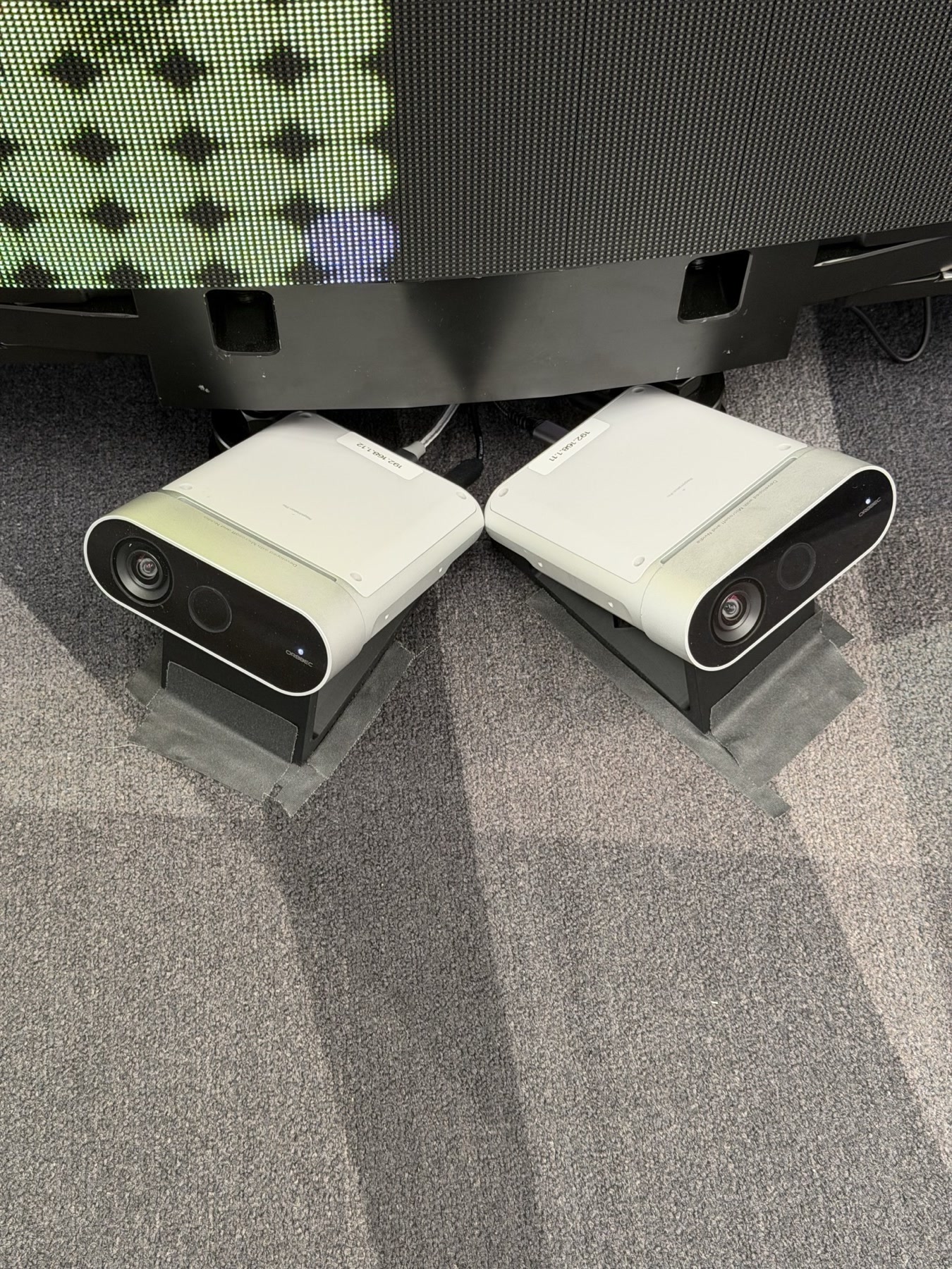 Two Orbbec Femto Mega depth sensors on 3D-printed mounts at the base of the curved LED wall, angled inward toward each other, with tape marks on the carpet around them
