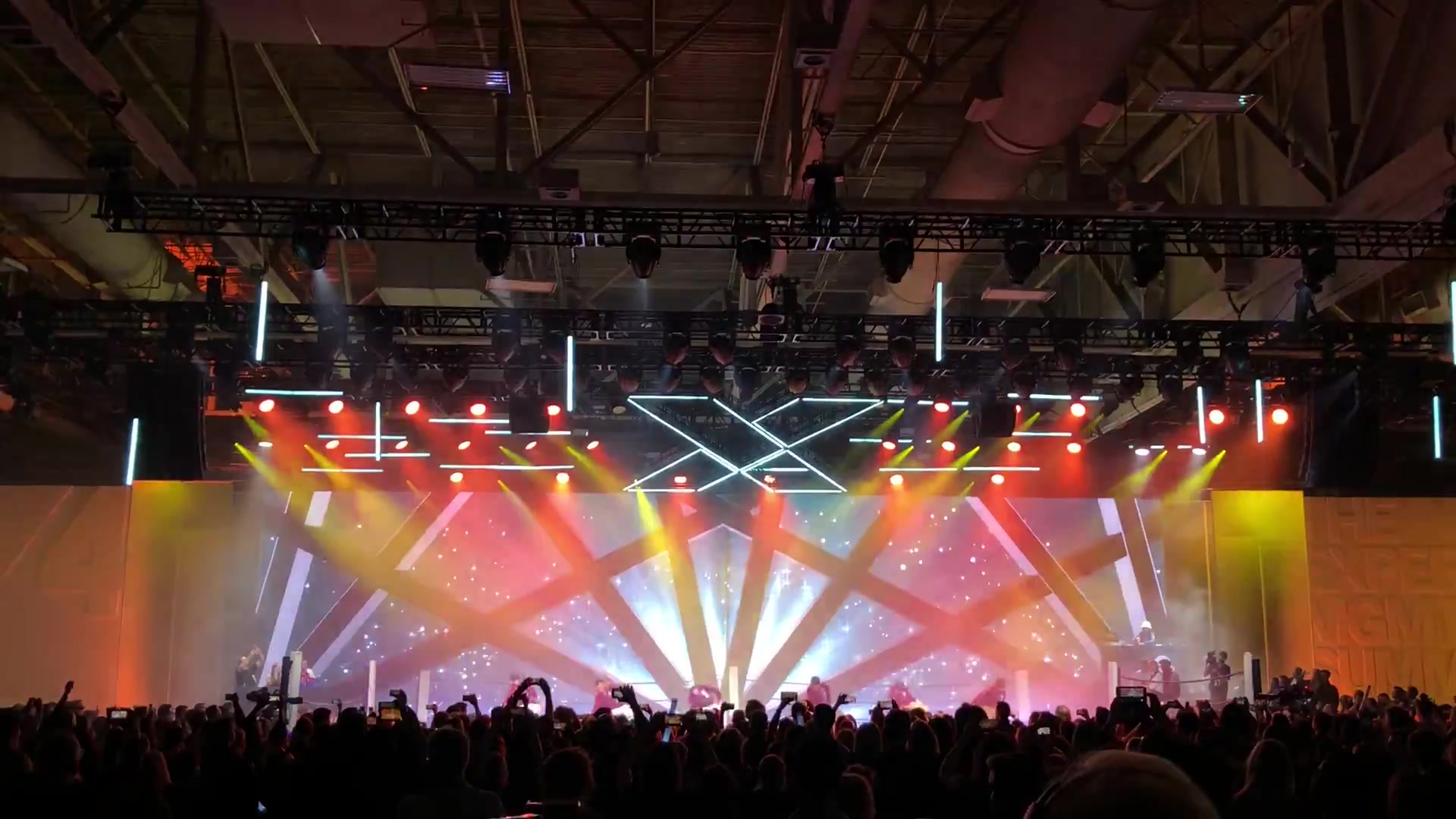 Symmetric fan of red, gold and white light beams radiating from a stage with an illuminated X4 logo truss overhead and performers in maroon jackets on stage, crowd with raised phones in the foreground